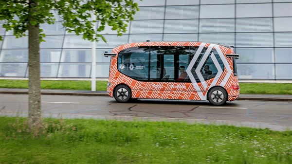 Avtonomni električni minibus Renault z oranžno-črnim kamuflažnim ovojem med testno vožnjo na mestni cesti ob zelenici.