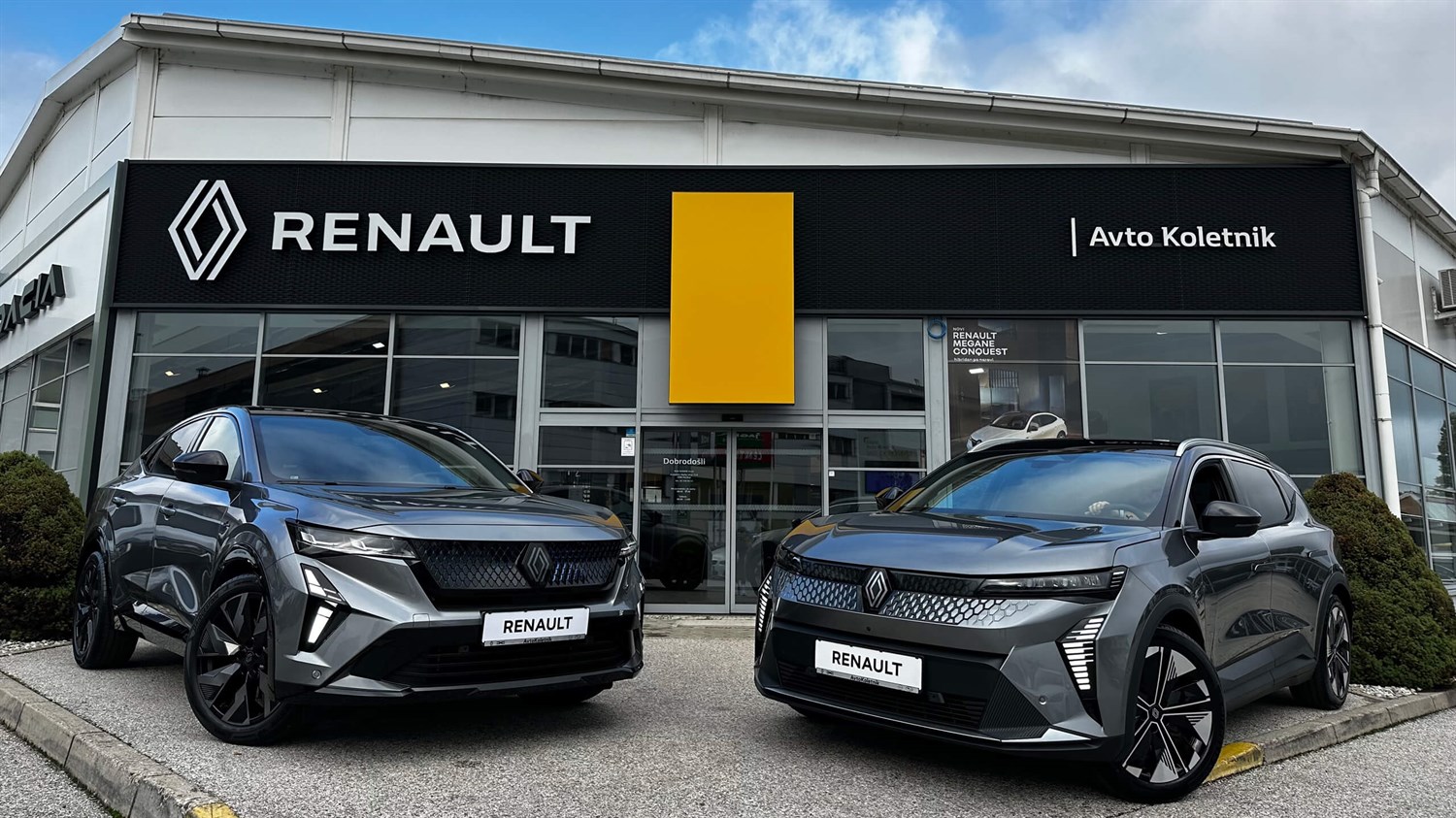 Dve sivi Renault vozili parkirani pred salonoma Avto Koletnik, nad vhodom velik napis Renault.