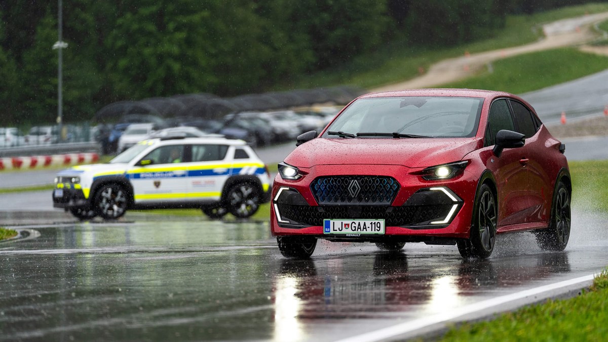 Rdeč avtomobil med vožnjo po mokrem voznem poligonu s policijskim vozilom v ozadju.