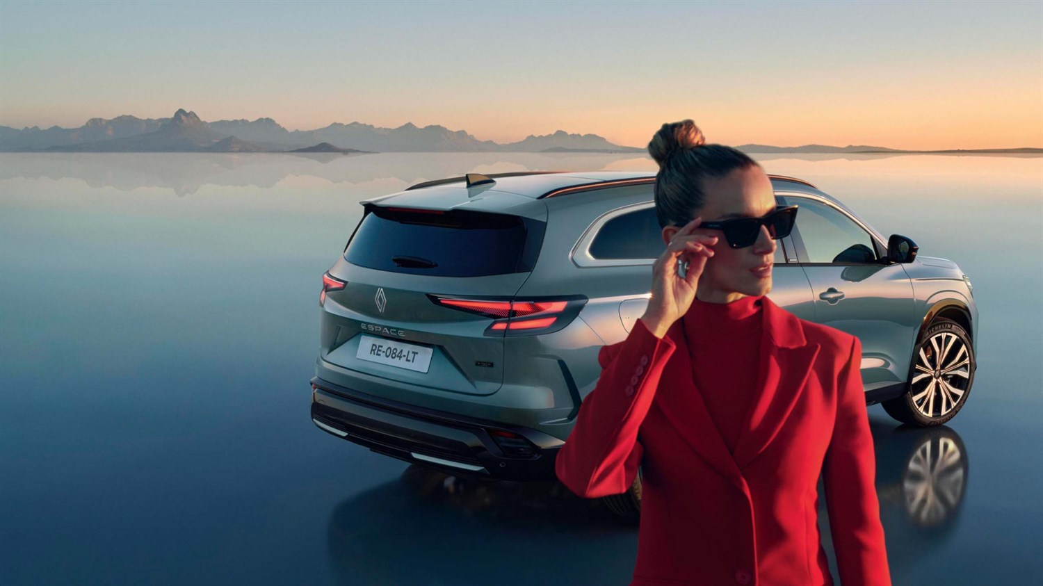 Sivi Renault Espace parkiran ob mirni vodni površini, v ospredju ženska v rdečem suknjiču.