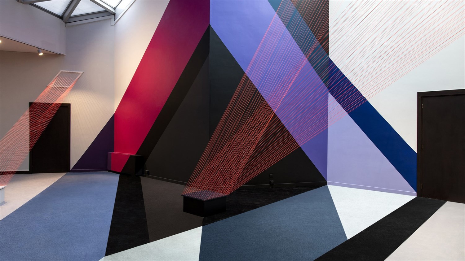 Umetniška instalacija z geometrijskimi oblikami in barvnimi linijami v galerijskem prostoru.