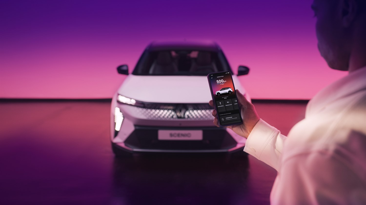 Moški drži pametni telefon z aplikacijo pred belim Renault Scenic, osvetljenim z vijolično-roza ozadjem.