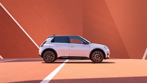 Bel manjši mestni avtomobil, fotografiran od strani na ozadju geometrijskih rdečkasto-rjavih ploskev.