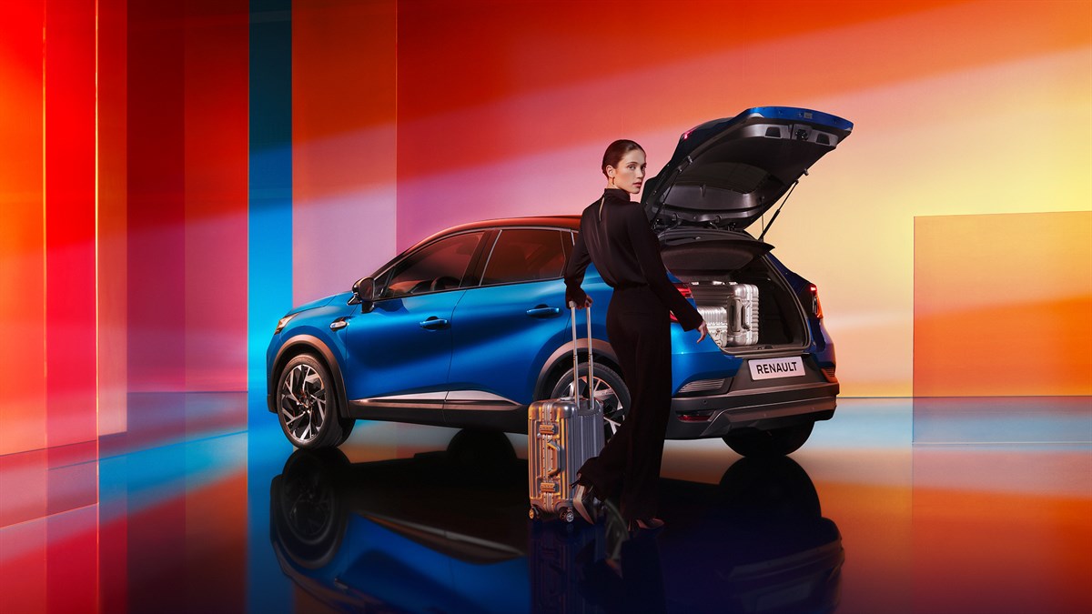 Na sliki je modri Captur z odprtim prtljažnikom, v katerem je kovček. Ob vozilu stoji elegantno oblečena ženska z ročno prtljago na kolescih, pripravljena na potovanje.
