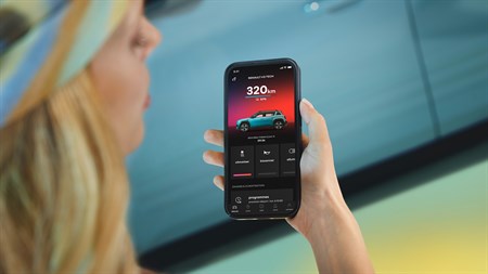 Ženska drži pametni telefon z aplikacijo Renault, ki prikazuje doseg električnega vozila 320 km, v ozadju je modro vozilo.
