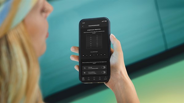 Svetlolasa ženska drži telefon z aplikacijo My Renault, na zaslonu prikazan urnik polnjenja z dnevno krivuljo in stikaloma za samodejno polnjenje.