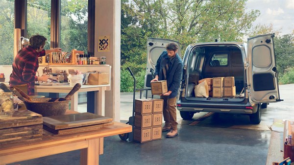 Renault Kangoo Van z odprtimi zadnjimi vrati pred skladiščem. Dva delavca nalagata tovor v prostoren tovorni del vozila.