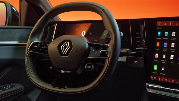 Volanski obroč vozila Renault Megane E-Tech Electric s poudarjenim logotipom Renaulta, digitalnim merilnikom hitrosti in velikim zaslonom na dotik z aplikacijami na sovoznikovi strani, ob oranžno-rdeči svetlobi iz okolice.