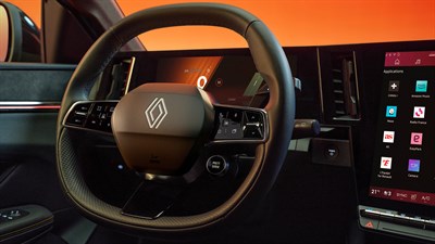 Volanski obroč vozila Renault Megane E-Tech Electric s poudarjenim logotipom Renaulta, digitalnim merilnikom hitrosti in velikim zaslonom na dotik z aplikacijami na sovoznikovi strani, ob oranžno-rdeči svetlobi iz okolice.