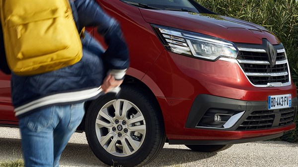 Sprednji del rdečega dostavnega vozila Renault z vidnim žarometom, prednjo masko in 43 cm (17'') platišči; v ospredju oseba z rumenim nahrbtnikom v gibanju. 
