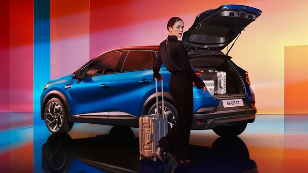 Modri Renault Captur z odprtim prtljažnikom, ob njem stoji ženska s kovčkom v barvno osvetljenem prostoru.